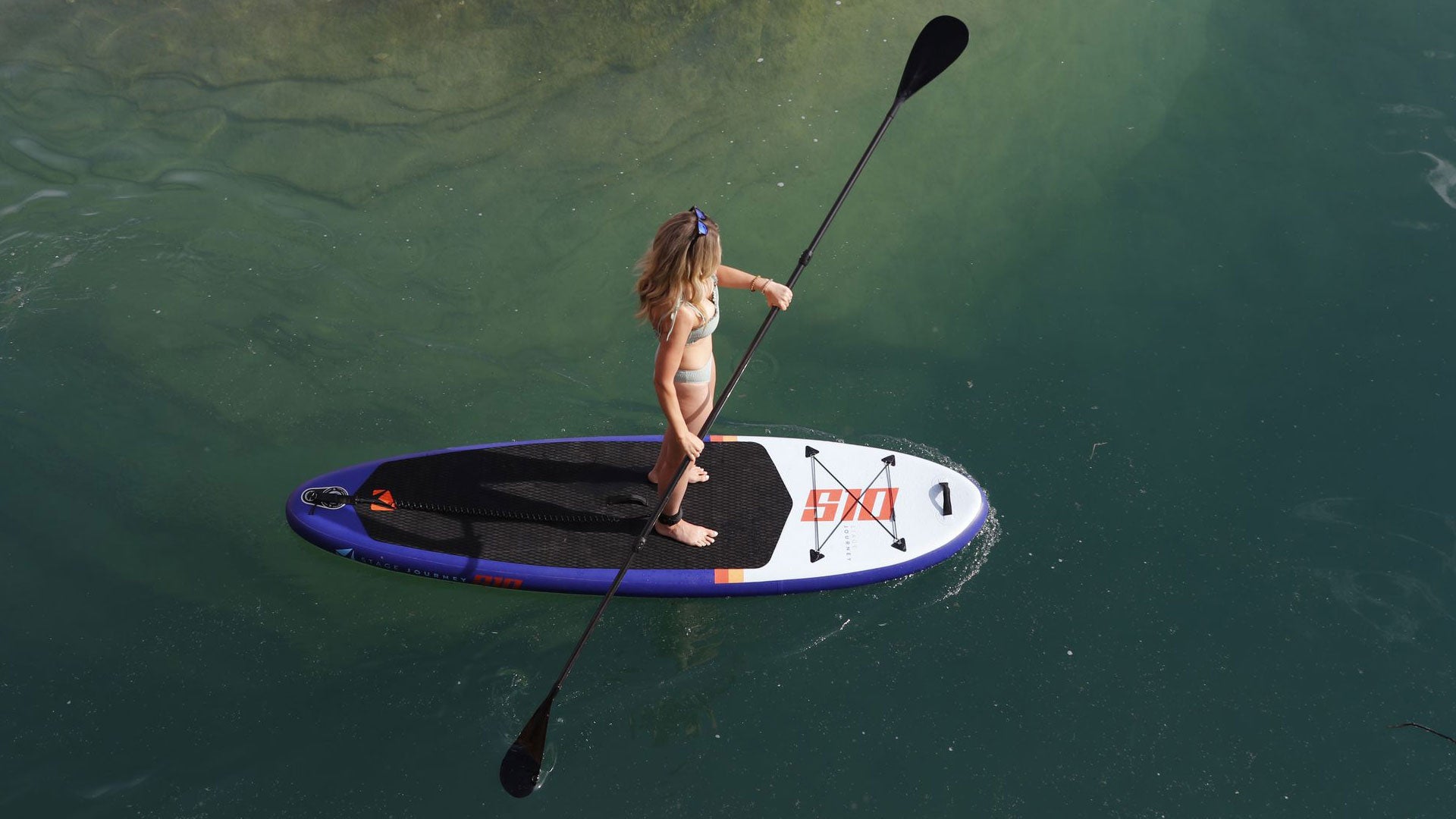 A woman paddling on a STAGE Inflatable Paddleboard and using the STAGE 2SIDE Double-Sided SUP Paddle in clear blue water, showcasing the board's stability and design.